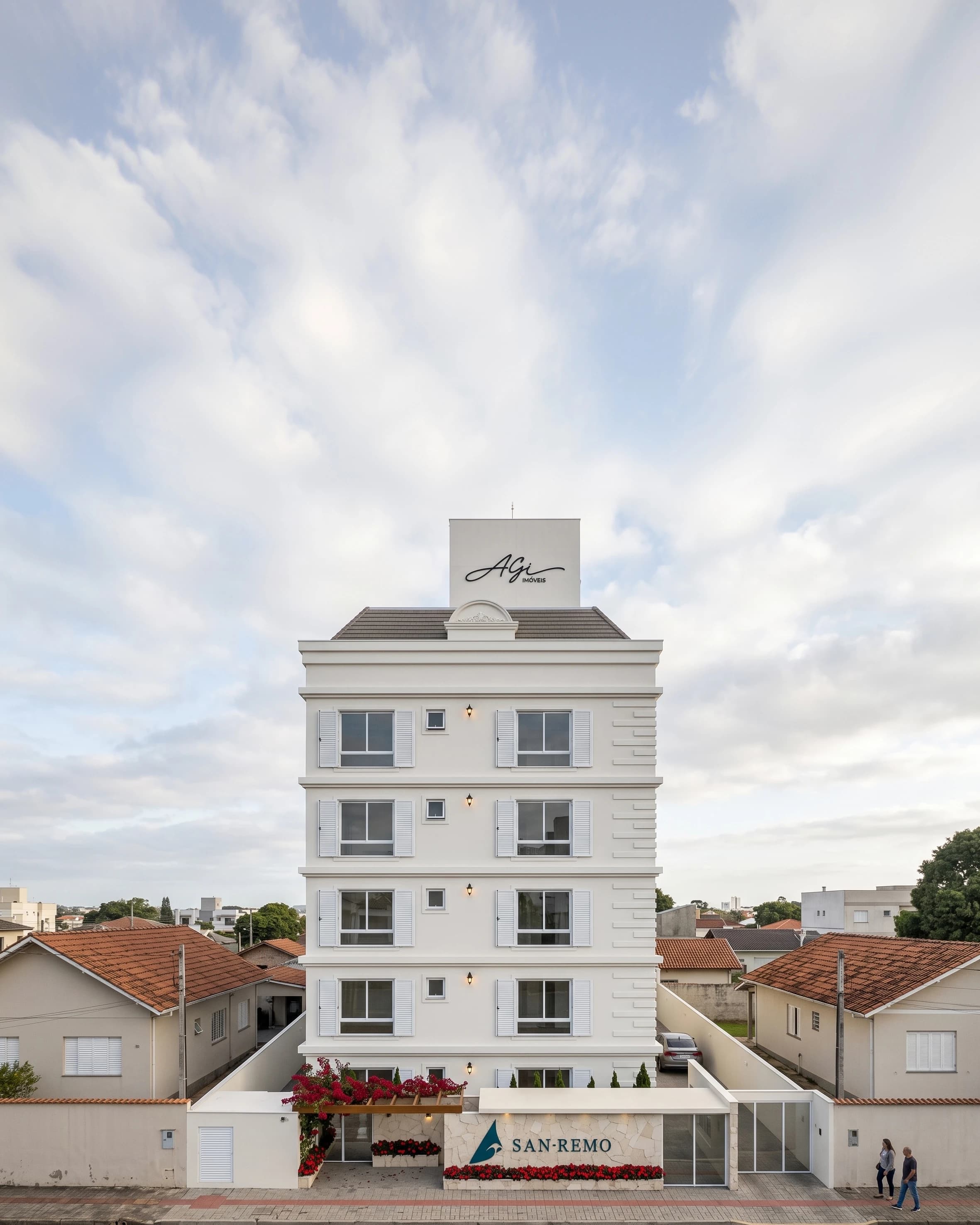 Fachada do San Remo de dia: edifício branco de cinco pavimentos com frontão, telhado aparente e venezianas, entre casas baixas.